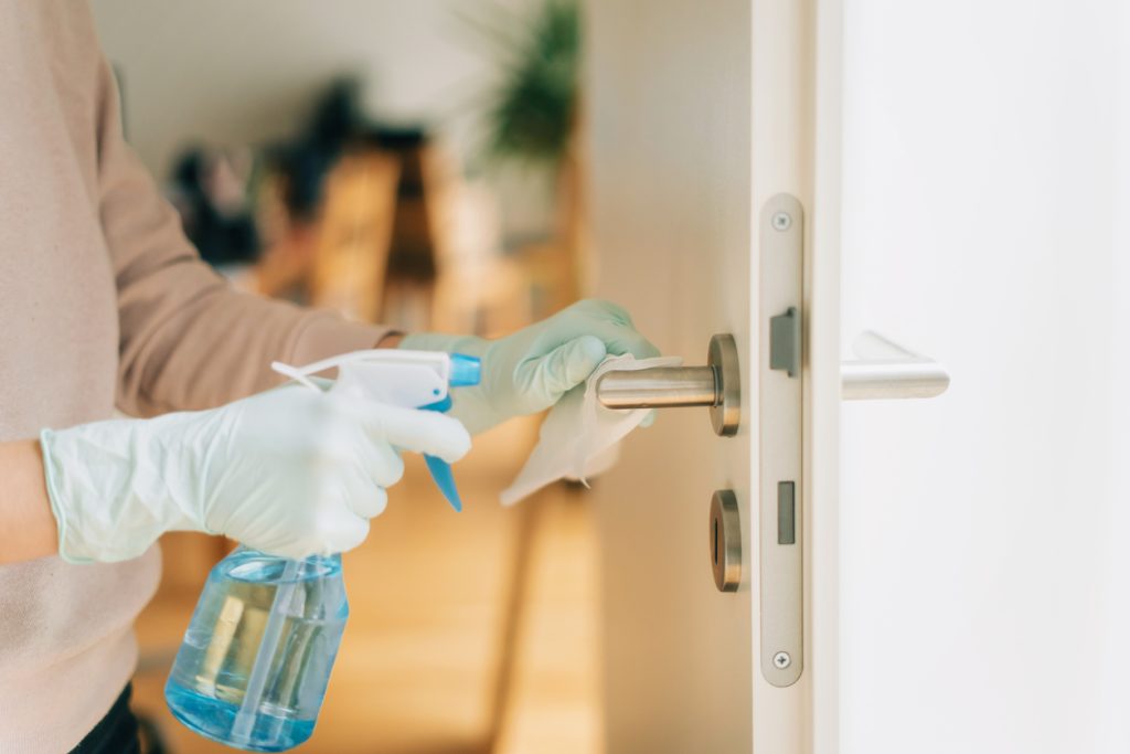 Woman in disposable gloves cleaning a door handle with a disinfection spray class=