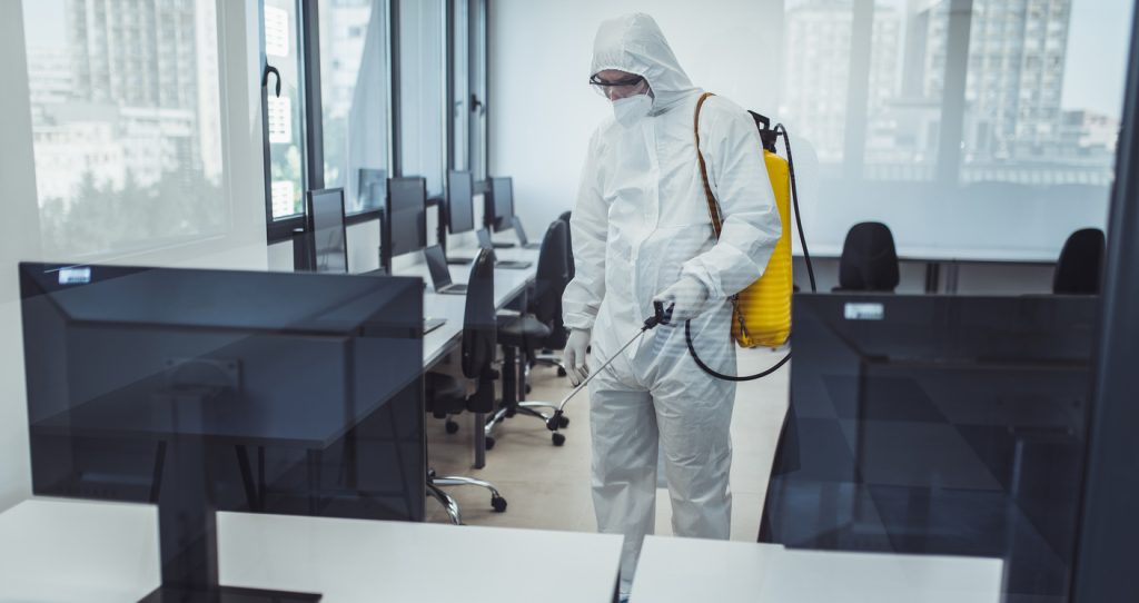 Office disinfection during COVID-19 pandemic. Man in protective suit and face mask spraying for disinfection in the office